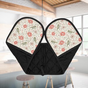May include: Black quilted oven mitts with a floral and bird pattern. The interior features a cream-colored background with red flowers, green leaves, and small birds. The mitts are designed with a quilted texture and a loop for hanging.