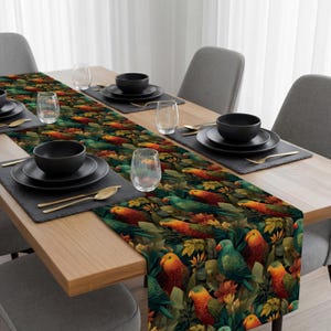 May include: A table runner featuring a vibrant parrot and foliage pattern in shades of green, orange, and blue. The runner is placed on a wooden table set with black plates, bowls, and gold-colored utensils. Clear glasses and gray chairs complete the dining setting.