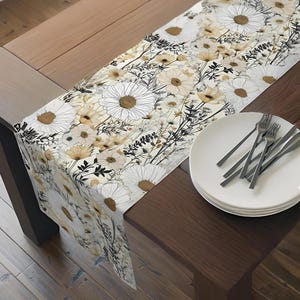 May include: A floral table runner featuring a daisy pattern in white, tan, and black. The runner is draped over a wooden table, with white plates and silverware on the right side. The design is a botanical theme.
