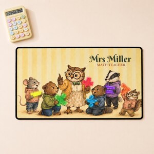 May include: A rectangular desk mat with a yellow striped background featuring an illustration of an owl teacher and animal students holding colorful math symbols. The text reads Mrs Miller MATH TEACHER. A light-colored calculator is positioned above.