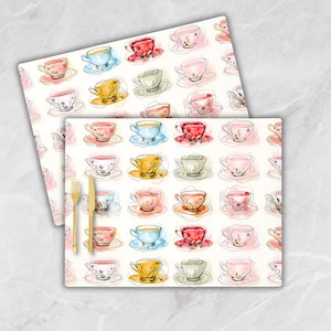 May include: A set of two white placemats with a repeating pattern of watercolor teacups in various colors, including pink, blue, yellow, and green. The teacups are outlined in black and have a delicate floral design.