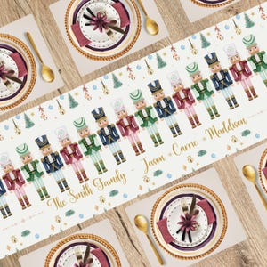 May include: A white table runner featuring a repeating pattern of toy nutcracker soldiers in various colors. The text "The Smith Family - Jason * Carrie * Maddison" is printed on the runner. The table setting includes plates, placemats, and gold-colored cutlery.