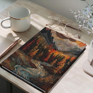 May include: A hardback journal with a landscape design featuring a river, trees, and mountains in shades of brown, orange, and blue. A white mug with a gold rim, a pen, and glasses are on the wooden surface.