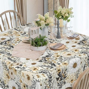 May include: A square white tablecloth with a floral pattern featuring white and yellow daisies, black stems, and brown leaves. The tablecloth is set on a wooden table with two chairs and a vase of white flowers.