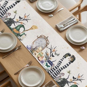 May include: A cream-colored Halloween-themed table runner with whimsical illustrations. The design includes witch legs, a green witch hat, a broom, a mirror with a spiderweb, and floral accents. The runner is placed on a wooden table with place settings.