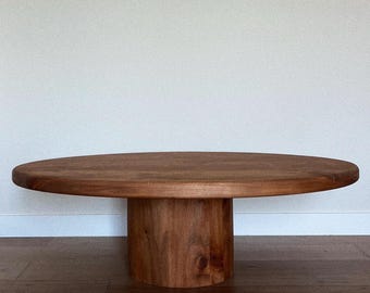 May include: A round wooden coffee table with a thick, oval-shaped top and a cylindrical base. The table is made of dark brown wood with a natural grain.