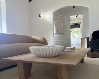 May include: A white ceramic bowl with a scalloped edge sits on a wooden coffee table with a light wood finish. The table has four legs and is in a living room with a beige couch and a large white vase with dried flowers.