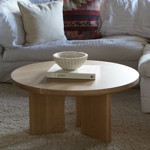May include: A round wooden coffee table with four legs. The table has a white book and a white ceramic bowl on top. The table is sitting on a white shag rug.