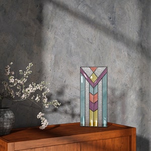 Könnte beinhalten: Ein rechteckiges Buntglasfenster mit geometrischem Design. Das Panel weist eine Vielzahl von Farben auf, darunter Lila, Gelb, Türkis und Pfirsich. Das Panel ist schwarz umrandet und steht auf einer Holzoberfläche.
