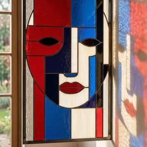 May include: A stained glass panel featuring an abstract face design. The artwork uses red, white, blue, and black geometric shapes to create the facial features. The panel is framed in black and mounted on a wall.