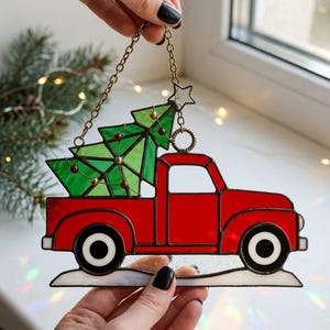 May include: A stained glass Christmas ornament featuring a red pickup truck carrying a green Christmas tree. The tree is decorated with small red and gold accents. The ornament is suspended by a chain and star.