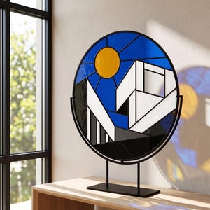Könnte beinhalten: Ein rundes Buntglas-Kunstwerk auf einem schwarzen Metallständer. Das Design zeigt einen blauen Himmel mit einer gelben Sonne, weiße geometrische Formen und schwarze und graue Akzente. Das Stück wird auf einem Holztisch ausgestellt.