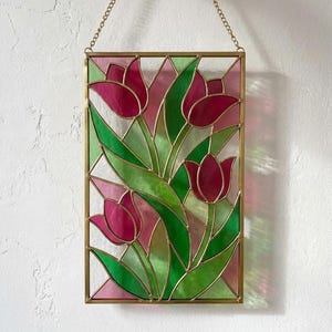 Könnte beinhalten: Buntglasfenster mit einem Blumendesign aus rosa Tulpen und grünen Blättern. Das Glas ist in einem goldfarbenen Metallrahmen gefasst und hängt an einer zarten Kette. Der Hintergrund ist ein hellrosa und grünes geometrisches Muster.