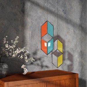 May include: A stained glass wall hanging with geometric shapes in orange, blue, yellow, and gray. The hanging is suspended from the ceiling with black string.