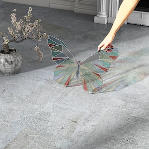 Könnte beinhalten: Ein bunter geometrischer Schmetterling aus Buntglas mit roten, grünen, blauen und gelben Akzenten. Der Schmetterling befindet sich auf einem grauen Betonboden.