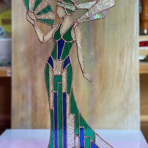 Puede incluir: Figura de vidriera de una mujer con un vestido largo, sosteniendo un abanico. El vestido es verde y azul con patrones geométricos, y la figura tiene alas y un tocado iridiscentes. Contornos color cobre definen el diseño.