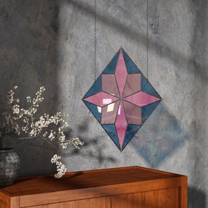 Könnte beinhalten: Ein rosafarbenes und blaues Bleiglasfenster in Form eines Sterns, das an einer Wand hängt. Das Fenster besteht aus kleinen Glasscheiben, die in einem geometrischen Muster angeordnet sind.
