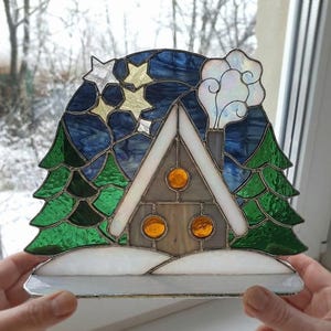 May include: Stained glass winter scene featuring a house with a snow-covered roof, flanked by green trees. The night sky is a deep blue with gold stars. White smoke rises from the chimney. The piece is held up by a person's hands.