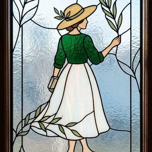 Puede incluir: Panel de vidriera que representa a una mujer con top verde, falda blanca y sombrero de paja, sosteniendo hojas. La obra presenta un tema de naturaleza con acentos de hojas y un fondo azul claro, enmarcado en madera oscura.