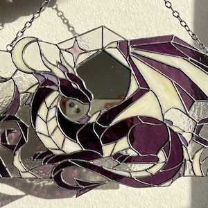 May include: Stained glass dragon wall hanging in shades of purple, white, and gray. The dragon is depicted in a geometric style with a mirror in the center. The piece is suspended by a silver chain.