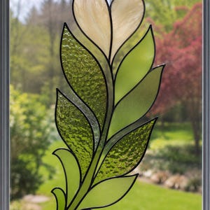 May include: A stained glass suncatcher shaped like a leafy branch with a heart-shaped top. The piece features various shades of green glass with smooth and textured finishes, accented by a pale cream heart, all held together by dark metal solder lines.