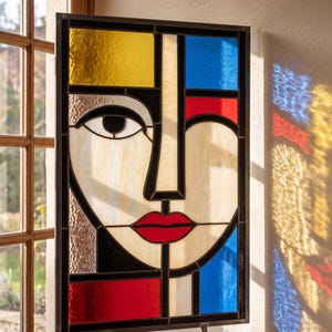 Könnte beinhalten: Abstrakte Glasmalerei mit einem stilisierten Gesicht. Das Kunstwerk hat ein geometrisches Design mit gelben, blauen, roten und weißen Glasabschnitten, die in Schwarz gerahmt sind. Das Gesicht hat eine rote Lippe und ein einzelnes Auge.