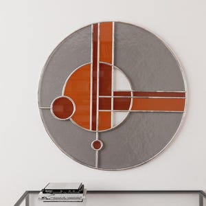 Könnte beinhalten: Ein rundes Wandbild aus Buntglas mit geometrischen Formen in Orange- und Grautönen.
