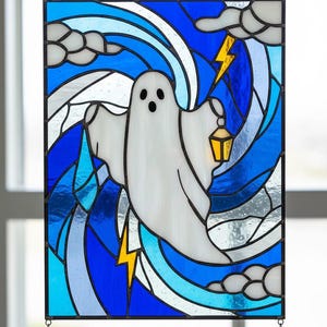Könnte beinhalten: Buntglasfenster mit einem weißen Geist, der eine Laterne hält, vor einem wirbelnden blau-weißen Hintergrund mit gelben Blitzen und Wolken. Das Panel ist schwarz gerahmt und hängt an einer Kette.