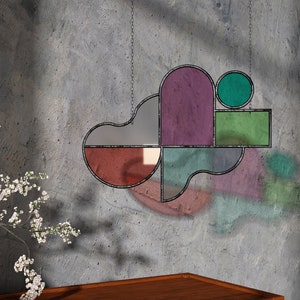 May include: A geometric stained glass window hanging with a purple, green, and red color scheme. The window is made of multiple pieces of glass that are joined together with black metal.