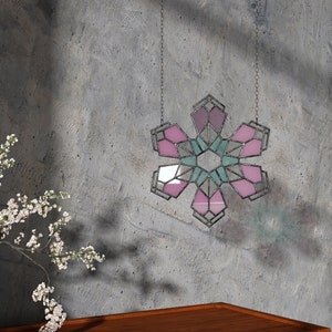 Könnte beinhalten: Ein Schneeflocken-Dekoration aus Buntglas hängt an einer Kette. Die Schneeflocke hat rosa, lila und türkisfarbene Glasscheiben, die von dunklem Metall umrahmt sind. Das Design ist geometrisch und symmetrisch, mit einer modernen Ästhetik.