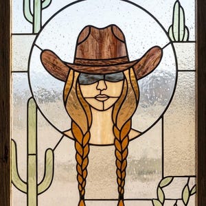 May include: Stained glass panel featuring a woman in a cowboy hat and sunglasses, with long braided hair. The design includes cacti and a circular frame, all set within a wooden frame. The color palette includes browns, tans, and greens.