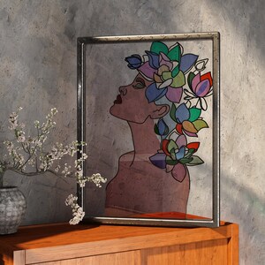 May include: Framed artwork featuring a woman's profile with a floral crown. The artwork uses a muted color palette with shades of brown, green, blue, and purple. The frame is a dark metal. The artwork is displayed on a wooden surface.
