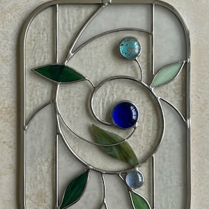 May include: Stained glass panel with a rectangular shape and a silver metal frame. The design features a swirling pattern with green and light green leaves, and three round glass beads in blue and turquoise colors. A small loop is at the top for hanging.