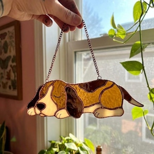 May include: Stained glass dog decoration in a sleeping pose, featuring brown, tan, and white colors. The dog is suspended by a copper-colored chain. The artwork is displayed in front of a window with natural light.