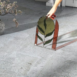 May include: A decorative stained glass panel featuring a stylized leaf design. The panel has a dark metal frame with a reddish-brown border and green and gold-toned glass. The design is held by a hand.