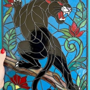Könnte beinhalten: Buntglasfenster mit einem schwarzen Panther mit roten Augen und offenem Maul, der auf einem Ast sitzt. Das Kunstwerk umfasst rote Blumen, grüne Blätter und einen blauen Hintergrund, alles in einem schwarzen Rahmen.