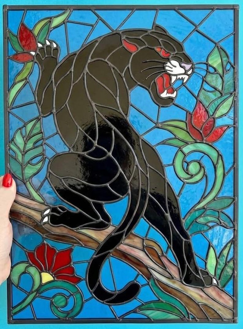 May include: Stained glass panel featuring a black panther with red eyes and open mouth, perched on a branch. The artwork includes red flowers, green leaves, and a blue background, all framed in a black border.