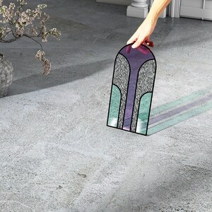 May include: A stained glass window panel with a geometric design in shades of purple, blue, and green. The panel is arched at the top and has a textured surface.