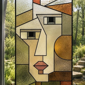 May include: A rectangular stained glass panel featuring an abstract human face composed of geometric shapes. The piece uses textured glass in shades of amber, olive green, cream, and brown, all outlined with black lead lines and suspended by a chain.