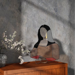 May include: A geometric mirror in the shape of the Mona Lisa, with black, gray, and red accents. The mirror is displayed on a wooden cabinet, with a vase of white flowers to the left.
