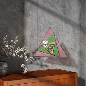 May include: A stained glass geometric triangle with pink, green, and yellow sections, suspended by chains. The artwork hangs against a textured gray wall, with a wooden cabinet and vase with flowers in the foreground. The design is a modern take on stained glass.