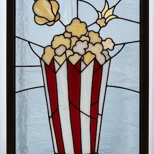 May include: Stained glass artwork featuring a popcorn-filled container with red and white stripes. The popcorn is yellow and white, with additional yellow accents. The artwork is framed in a dark brown wooden frame.
