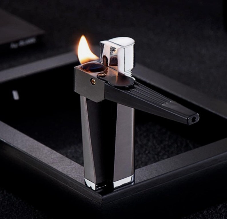 May include: A black and silver lighter with a flame coming out of the top. The lighter has a long, thin nozzle and a rectangular body.