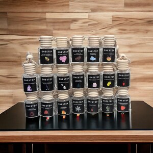 May include: A collection of 18 small glass bottles with wooden caps and colorful labels. Each bottle has a different scent and design. The bottles are arranged in three rows of six. The bottles are on a wooden surface.