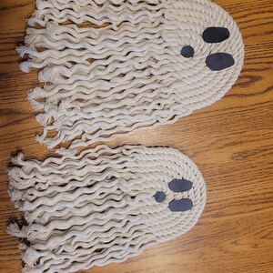 May include: Two white rope ghosts with black felt eyes. The ghosts are hanging on a wooden surface.