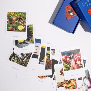 May include: A collection of oracle cards with various images and text, scattered on a white surface. The cards feature colorful illustrations and phrases. A blue box with the text "Lost & Found Oracle" is also present.