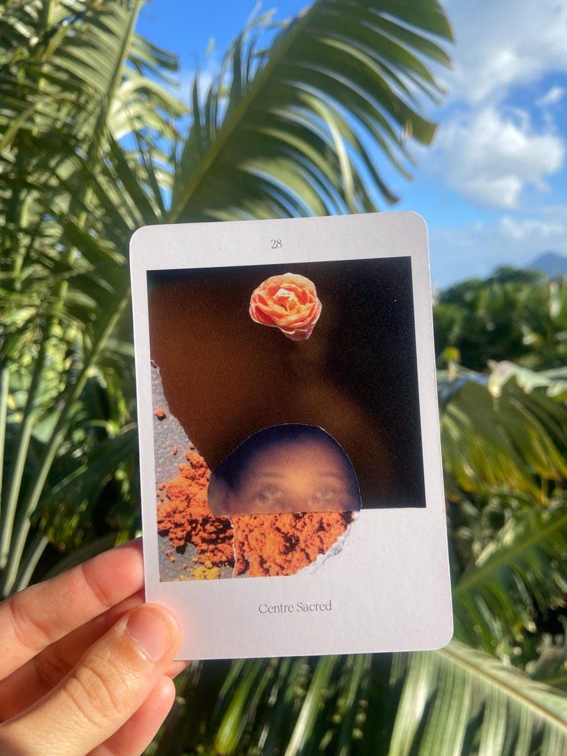 May include: A tarot card with a torn edge, featuring a rose and a partial face. The card has the number 28 at the top and the words "Centre Sacred" at the bottom. The background includes palm leaves and a blue sky.