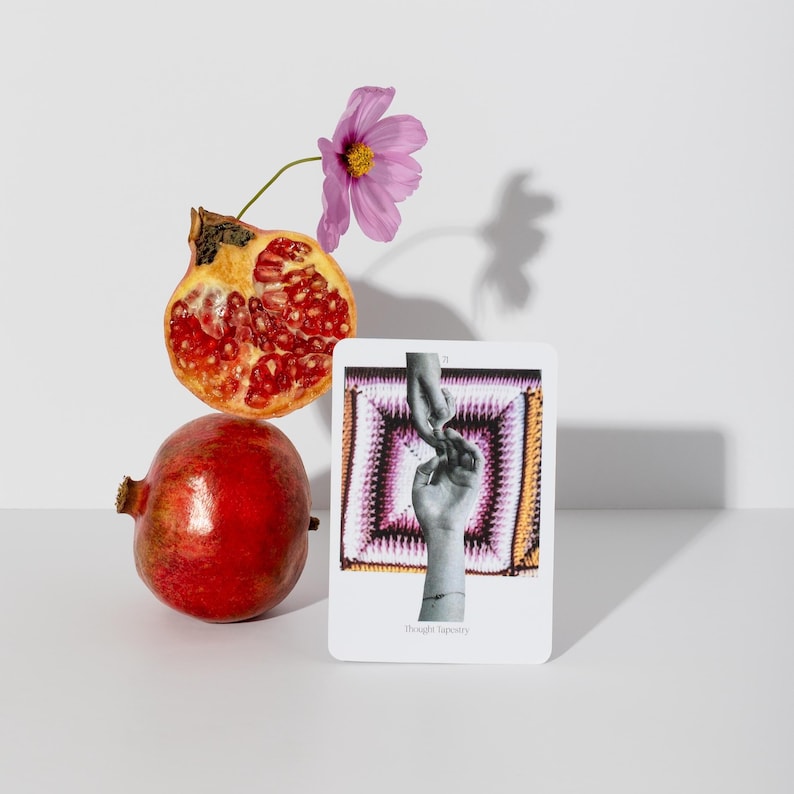 May include: A still life arrangement featuring a whole pomegranate and a halved pomegranate with visible seeds. A pink flower and a card with a hand-holding image are also present. The card has the text "Thought Tapestry".