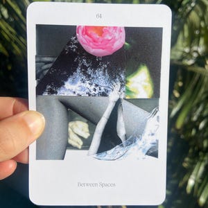 May include: A white card with the number 64 at the top and the words "Between Spaces" at the bottom. The card features a collage with a pink flower, grayscale images of body parts, and abstract shapes. The card is held by a hand.
