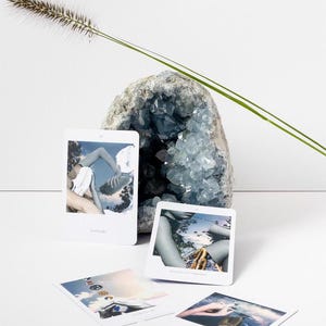 May include: A blue geode crystal formation with several small, white rectangular cards featuring images. A tall, thin green plant stem and a fluffy brown plant head are also present. The cards have text.